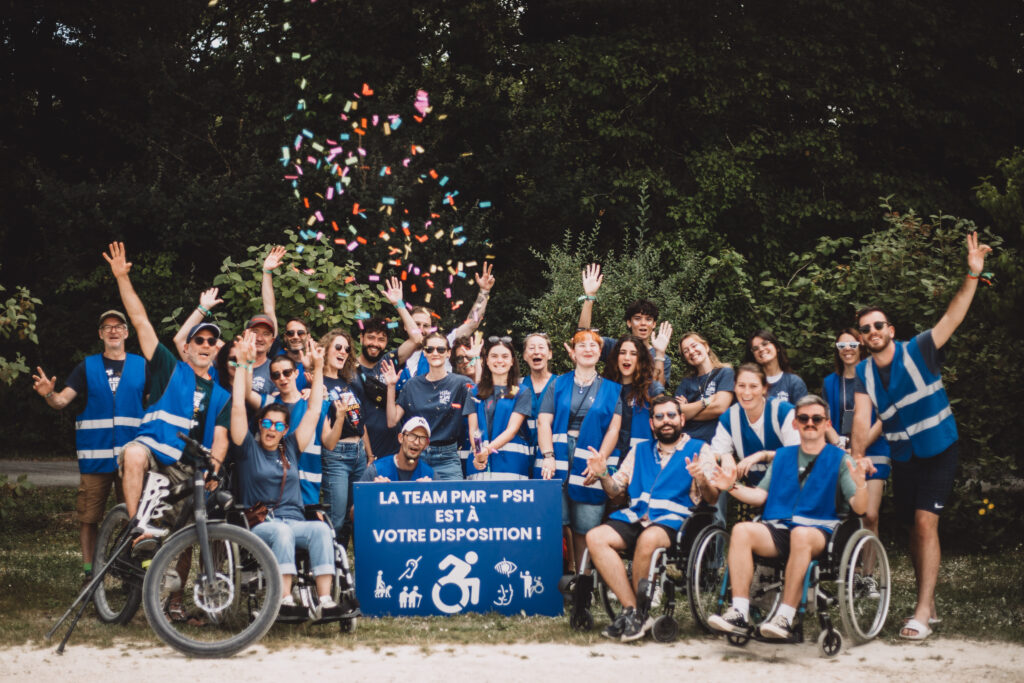 Une photo de groupe des bénévoles avec les mains en l'air, certain son assis sur des fauteuils roulant et ont tous des gilets réfléchissant de couleur bleu. Une pancarte avec écrit dessus "la team PMR - PSH est à votre disposition !"