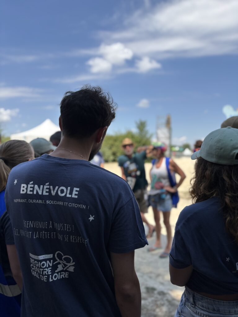 Un bénévole avec un t-shirt avec écrit bénévole qui est de dos et écoute les instructions d'une personne en face de lui.